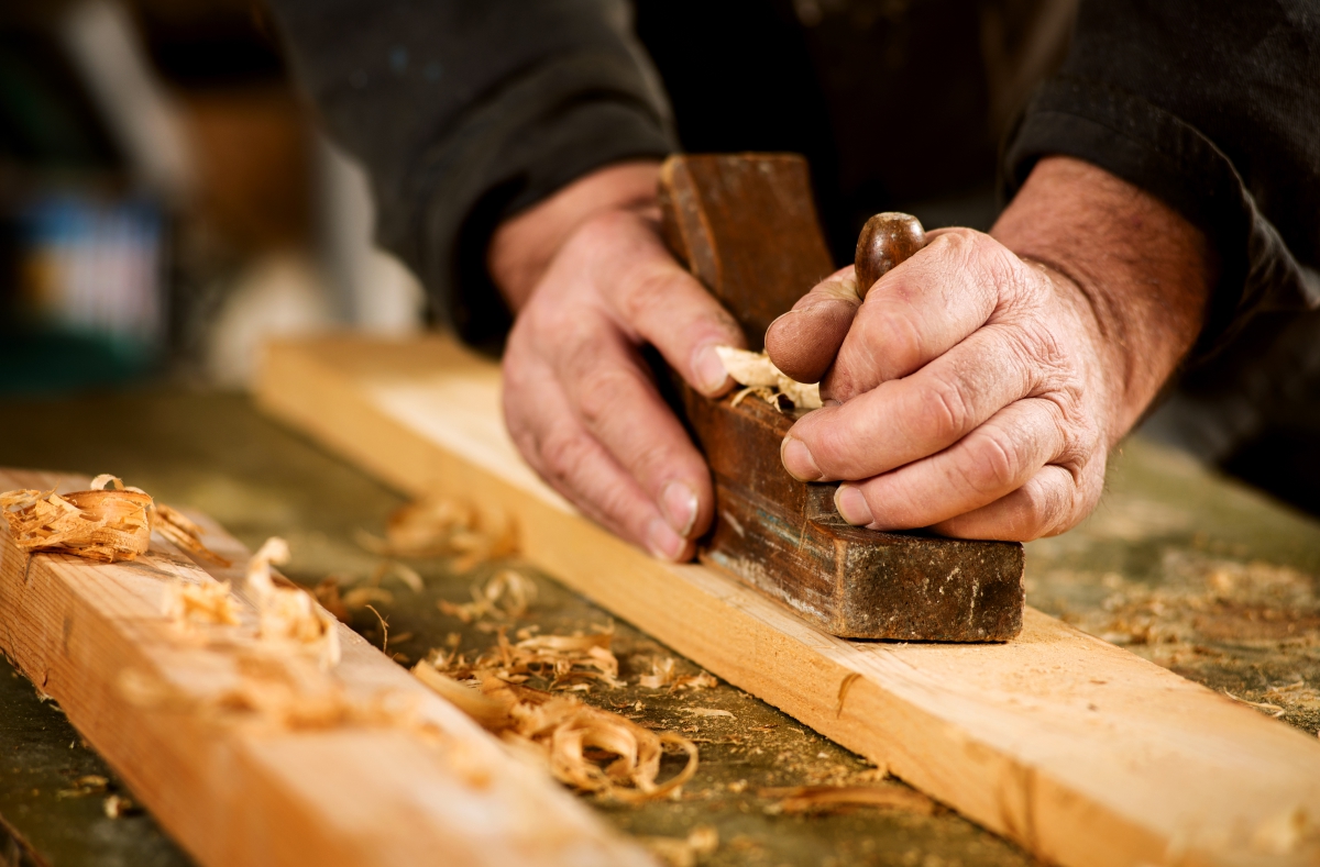 Skilled carpenter using a handheld plane to smooth and level the surface of a plank of hardwood, close up view of his hands, the tool and wood shavings.